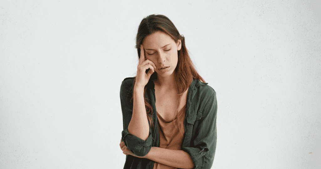 A woman stands against a white wall with her eyes closed and a hand pressed to her temple, portraying the deep internal exhaustion caused by invisible social pressures.