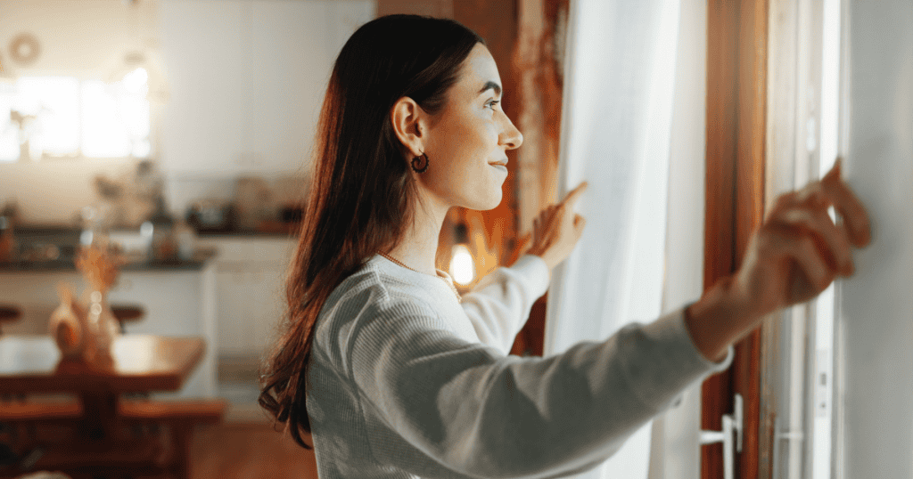 A woman pulls back white curtains to let in the morning light, visualizing a cautious emergence into her own environment.