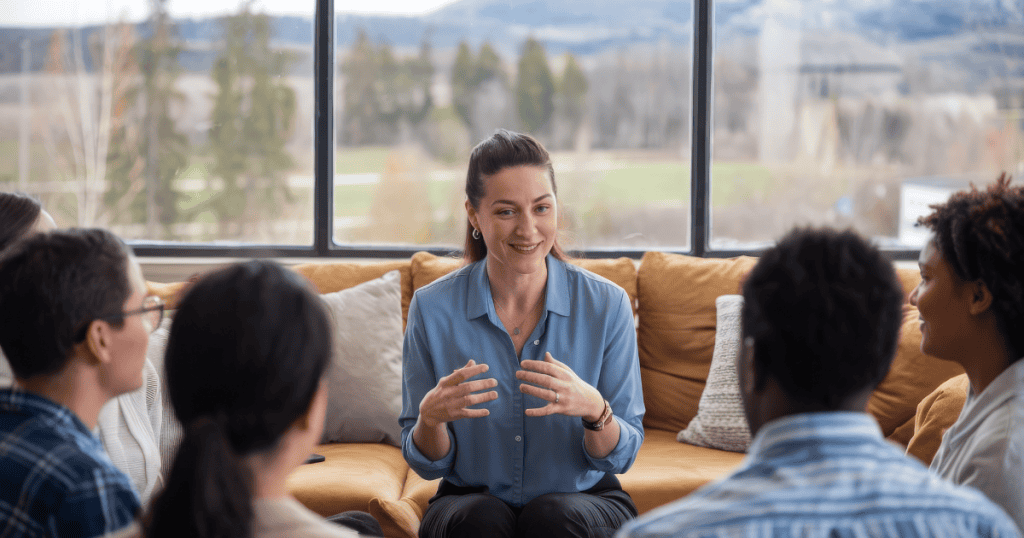 A woman smiles warmly while gesturing to a seated group, her poised demeanor masking a total lack of personal responsibility.