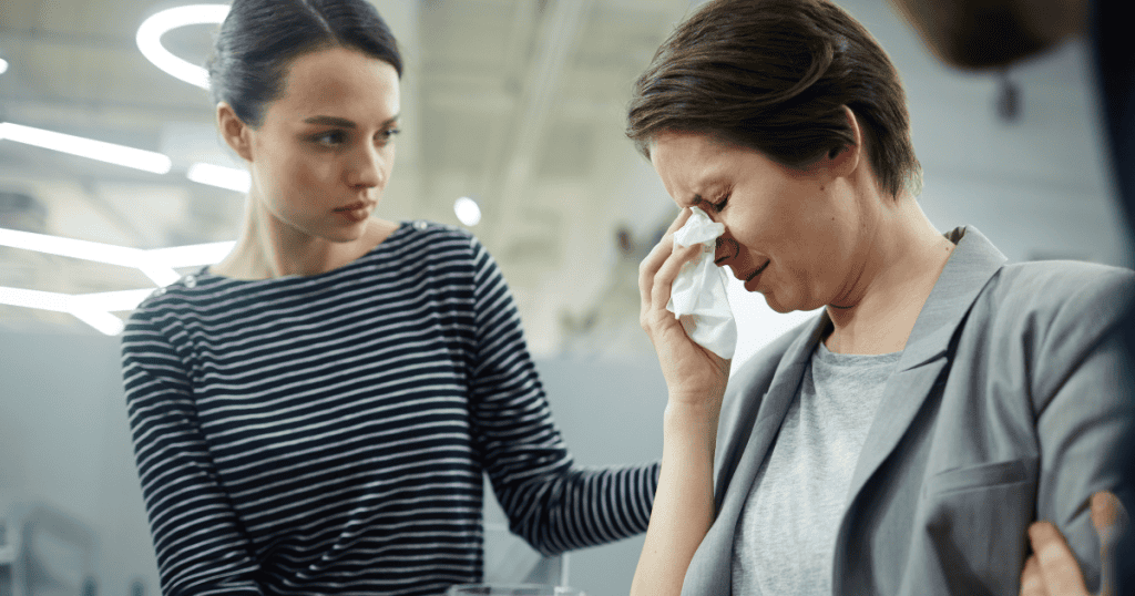 A woman weeps into a tissue while another person watches with a skeptical expression, highlighting a moment of intense, inward-focused sorrow.