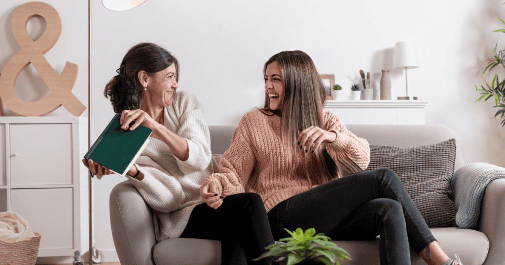 Two women sit close together on a couch laughing and sharing a book, representing the balanced, joyful energy of a truly reciprocal connection.