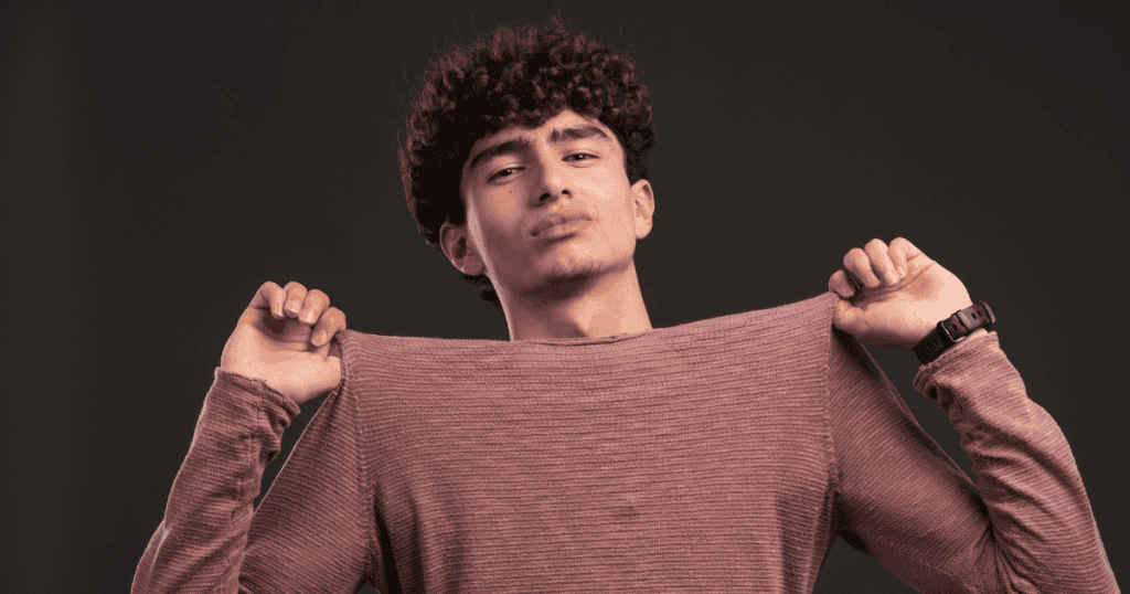 A teenage boy with curly hair pulls at the collar of his pink sweater with a defiant gaze, illustrating the transition into using appearance as a defensive social shield.