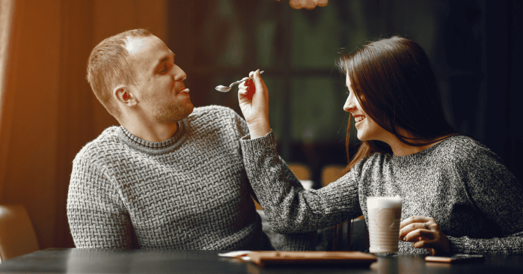A man and woman sit at a café table laughing while she feeds him a spoonful of dessert, portraying a moment of domestic comfort that begins to be tested by the weight of genuine connection.