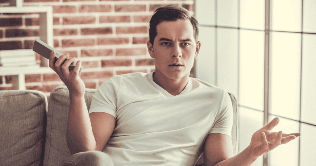 A man sits on a couch holding a smartphone with an expression of confusion and frustration, capturing the growing agitation when familiar sources of validation start to slip away.