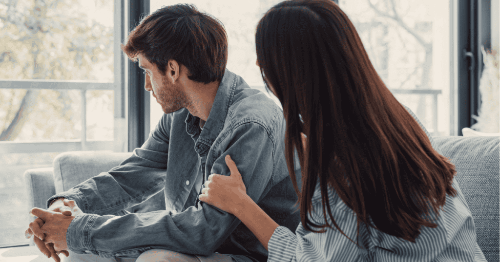A woman places a hand on the shoulder of a man who is looking away toward a window, capturing the moment an attempt at connection is met with a cold, deliberate withdrawal.