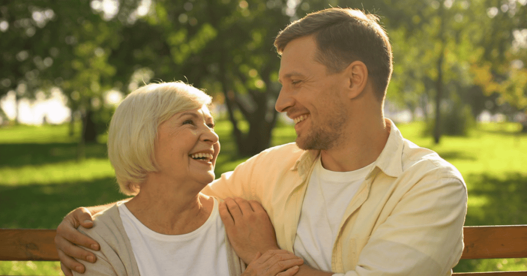 An older blonde woman and a younger man sit on a wooden bench laughing together in a sun-drenched park, capturing an idealized display of deep familial devotion and warmth.