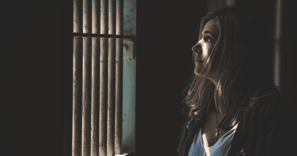 A young woman stands in a dim room looking toward the light coming through a set of vertical metal bars, symbolizing the realization of being confined within a repetitive, invisible structure.