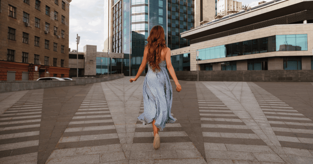 A woman in a long flowing dress walks directly away from the camera toward a wide city plaza, illustrating a definitive path toward independence taken without looking back.