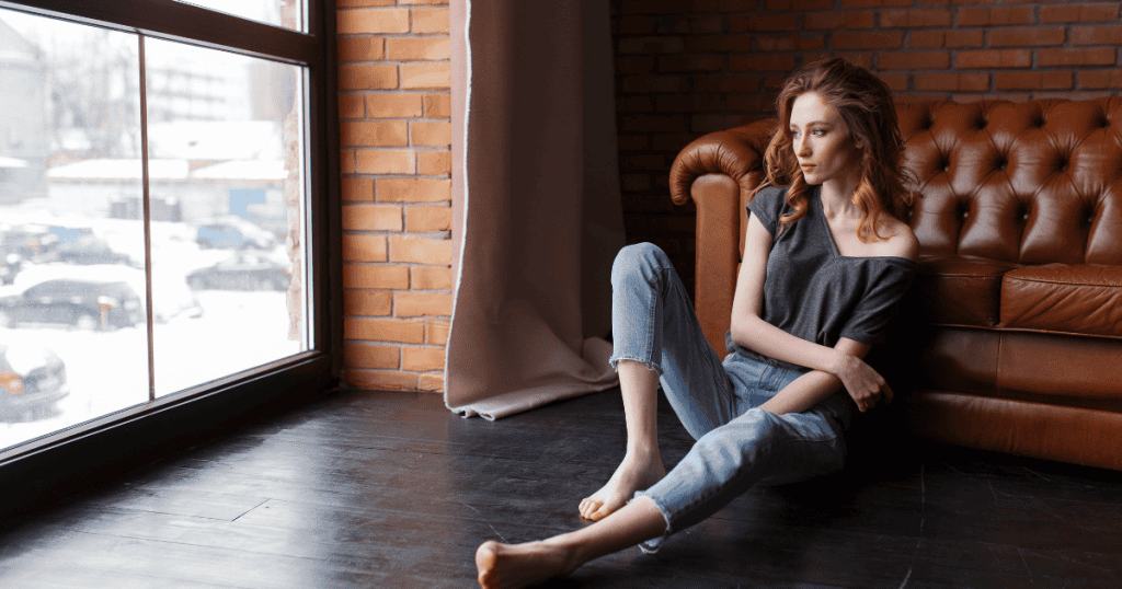A woman sits on the floor by a large window in a brick-walled room, reflecting a quiet sense of isolation when answers no longer come.