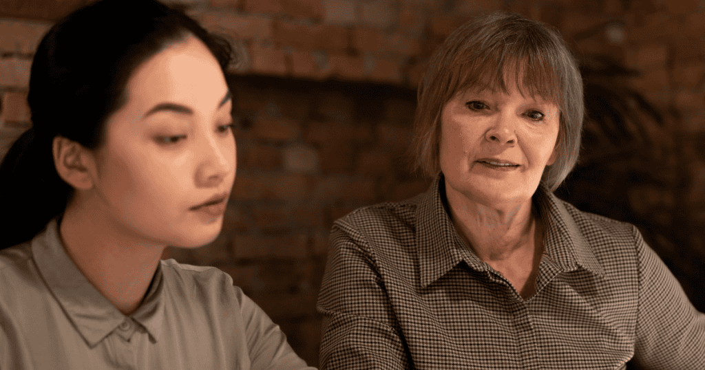 An older woman looks toward a younger woman who is looking down, highlighting an unspoken distance where roles have suddenly shifted.