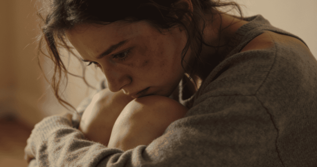 A young woman sits huddled on the floor with her head resting on her knees, visibly burdened as if you carry a low-level, persistent sadness.