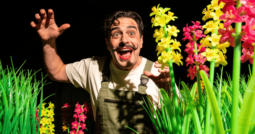 A man with an exaggerated mustache and a wide, intense grin poses dramatically behind tall garden flowers, showcasing a highly theatrical performance of a classic weekend pastime.