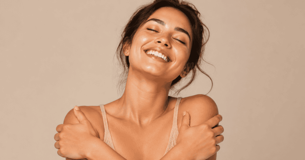 A woman beams with an open smile while closing her eyes and wrapping her arms around her own shoulders, beautifully demonstrating how coming back to yourself is the real breakthrough.