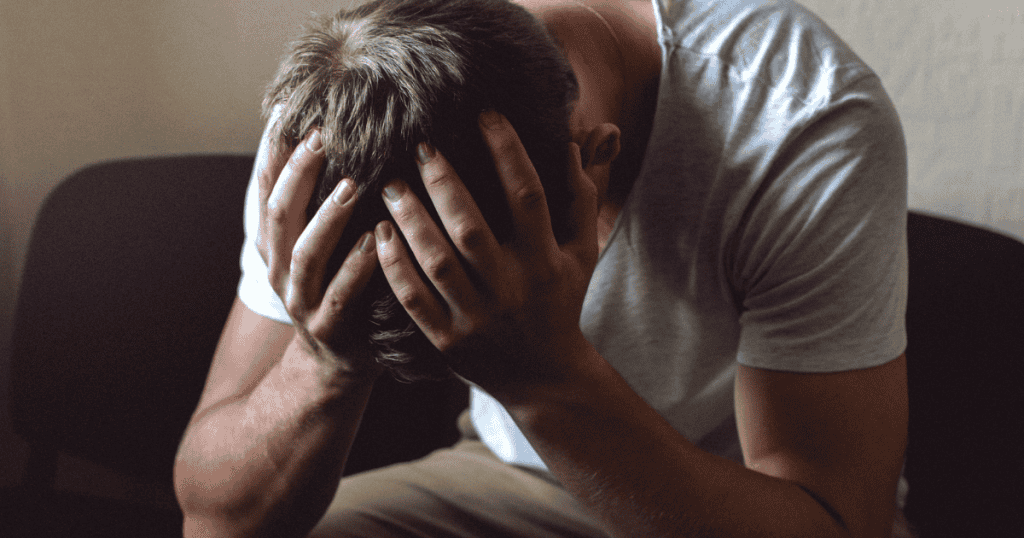 A distressed man sits on a dark couch with his head buried tightly in his hands, reflecting a profound sense of psychological collapse and final desperation.