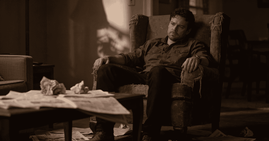 A somber man sits slumped in a heavily worn, tattered armchair inside a dimly lit living room, completely surrounded by a quiet isolation that has slowly accumulated over time.