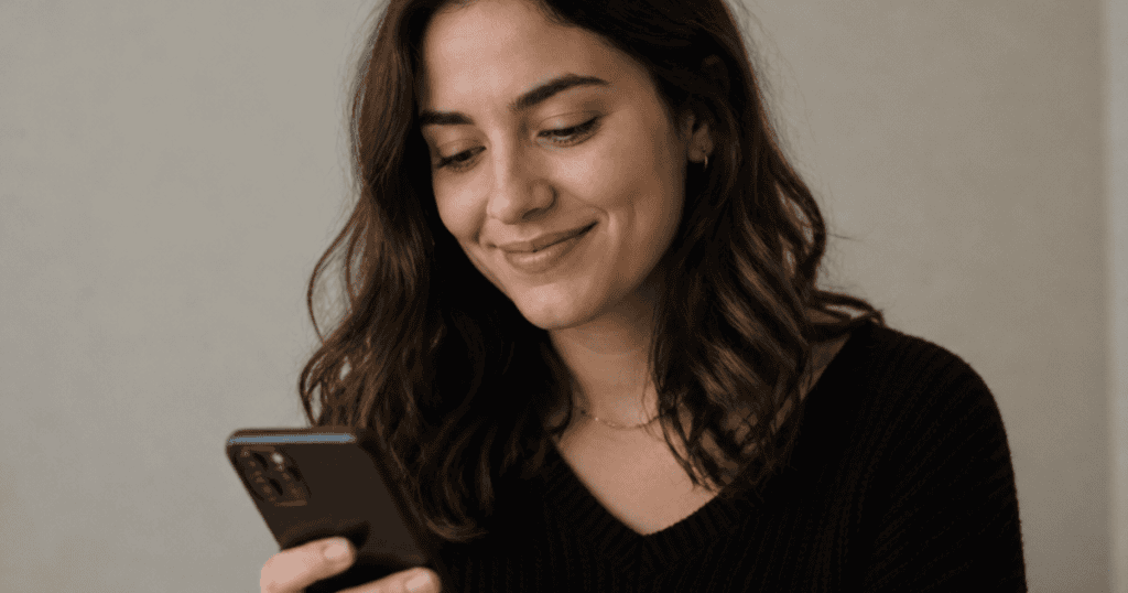 A young woman looks down at her smartphone with a serene, genuine smile, embodying the peaceful liberation of anchoring one's thoughts completely outside of someone else’s orbit.