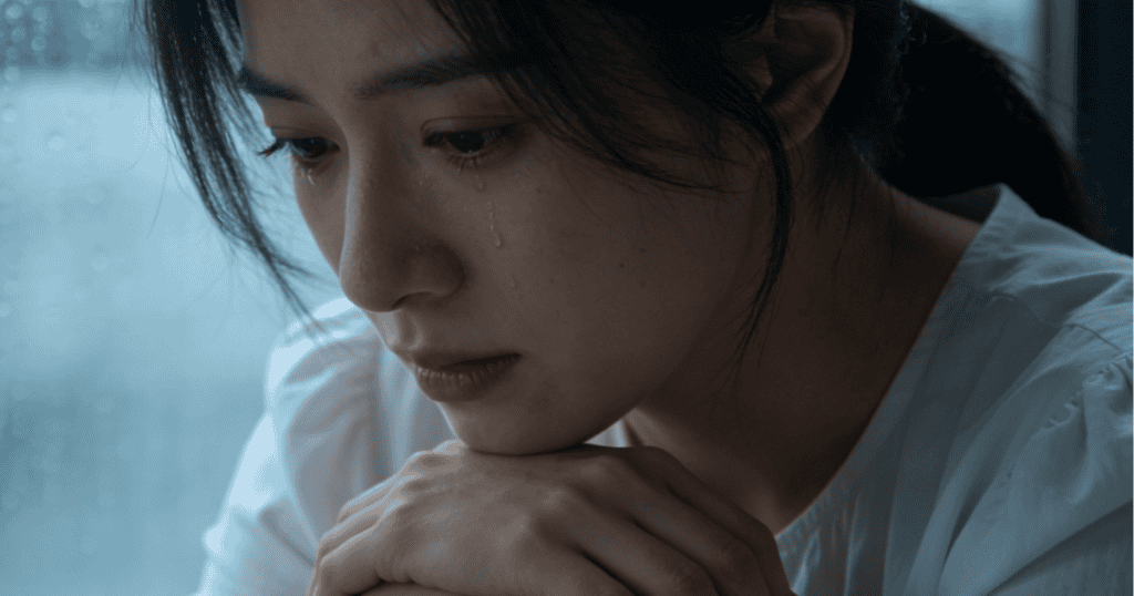 A young woman looks down with a somber expression as tears stream down her cheek while resting her chin on her hands, embodying a heavy state of emotional isolation that goes completely unseen by the outside world.