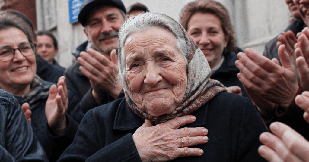 An elderly woman in a patterned headscarf presses her hands to her chest with a modest, grateful smile while an affectionate crowd applauds behind her, illustrating an impeccable public reputation built on perceived humility.