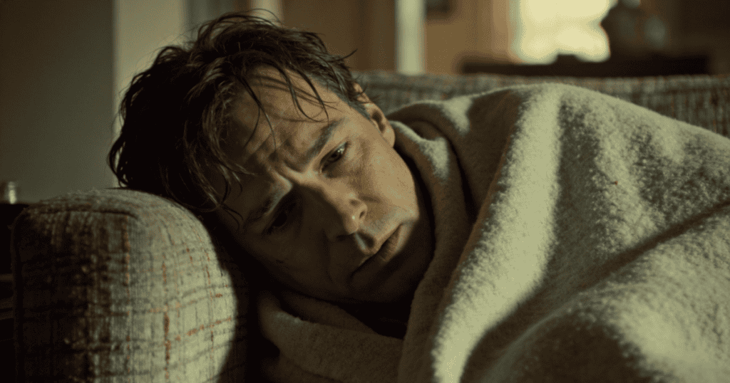 A pale, sickly man lies dejectedly on a couch wrapped in a heavy blanket, staring blankly ahead into an empty room where no one remains to answer his silent distress.