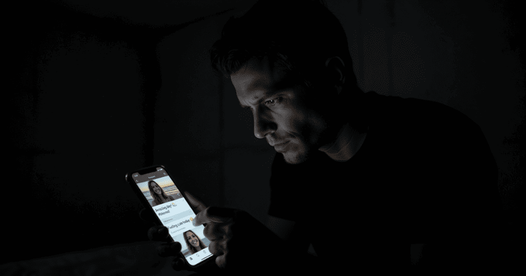 A man sits in a dark room staring intently at a glowing smartphone screen, as if searching for a external justification to shift the blame for his own choices.