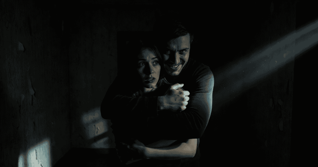 A sinister man smiles creepily while holding a frightened woman tightly from behind in a dark room, symbolizing the unsettling way some thrive on another's suffering.