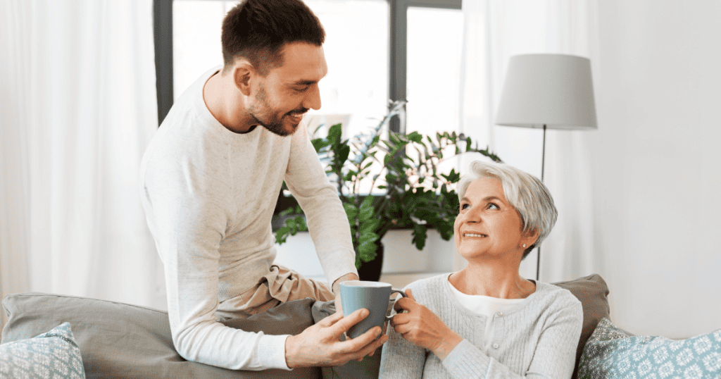 A man leans forward to hand a mug of tea to a smiling senior woman sitting on a sofa, depicting an overly attentive gesture designed to leave a flawless first impression.