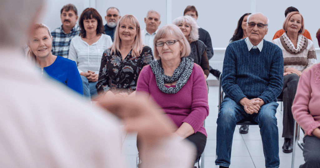 An audience of diverse adults sits listening attentively to an unseen speaker in the foreground, capturing the curated social facade built to project widespread approval and evade criticism.