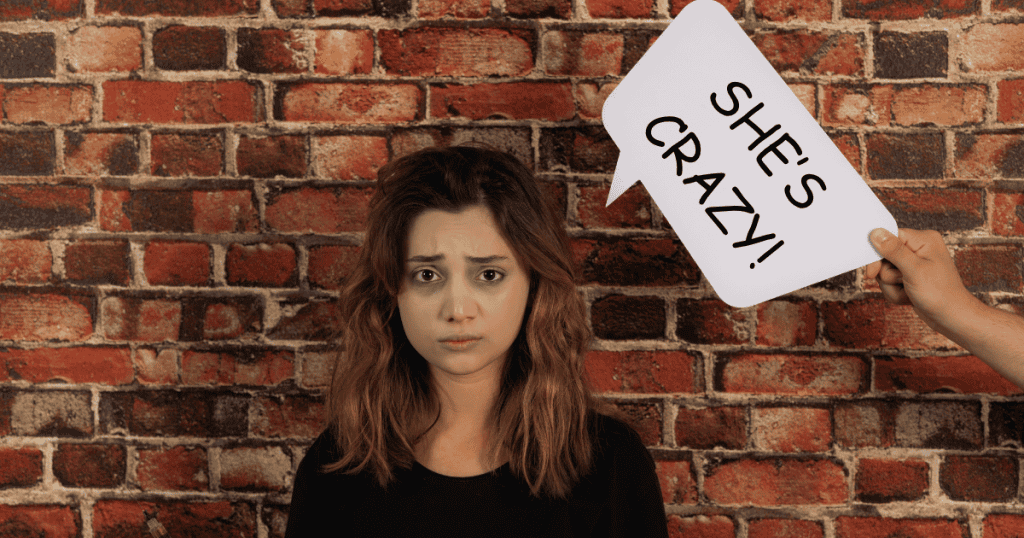A hand holds up a speech bubble reading "SHE'S CRAZY!" next to a wide-eyed, exhausted woman against a brick wall, symbolizing the weaponization of false narratives to discredit emotional distress.