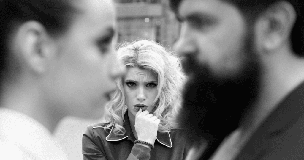 A worried blonde woman bites her nail in the background between two blurred faces in sharp black and white, illustrating the quiet tension felt around people who might uncover hidden intentions.