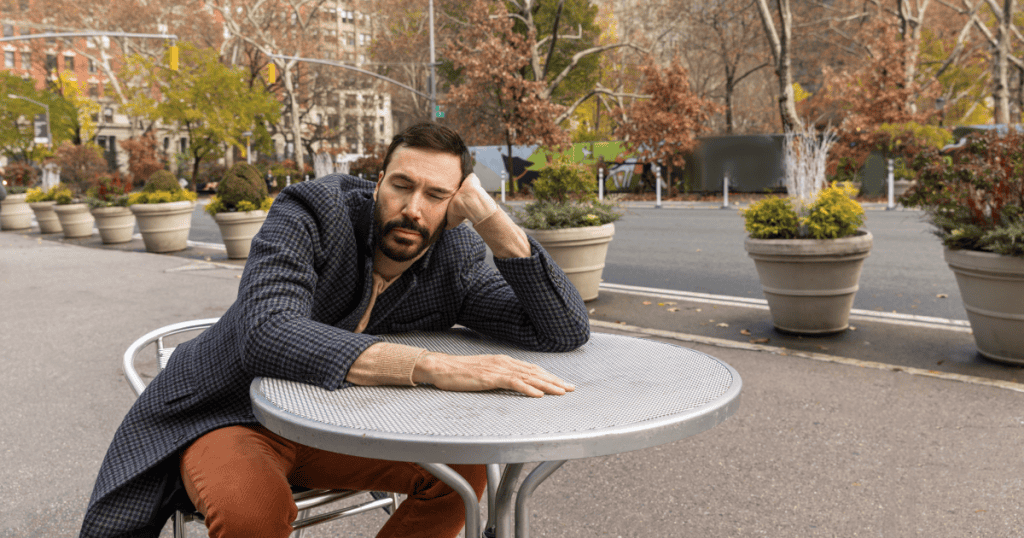 A man slumped alone at an outdoor table with a defeated, hollow expression, capturing the total collapse a narcissist spirals into after a humiliating defeat triggers their most dangerous version.