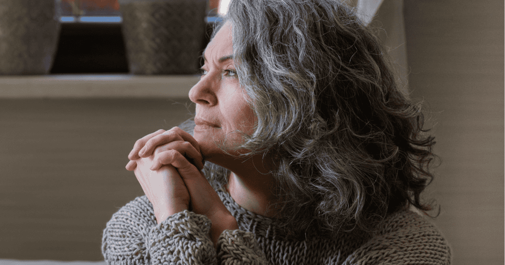 A middle-aged woman with silver hair clasps her hands beneath her chin while looking out a window, embodying the deep introspection and quiet solitude that self-absorbed individuals constantly avoid.