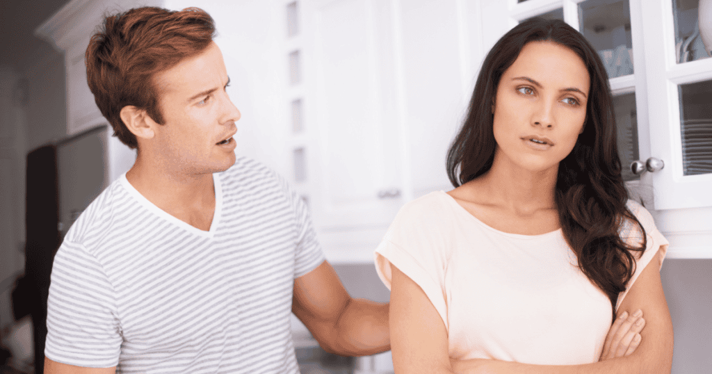 A woman stands with crossed arms turning away from a man speaking defensively in a kitchen, portraying the instinctive boundary set against someone probing for leverage.