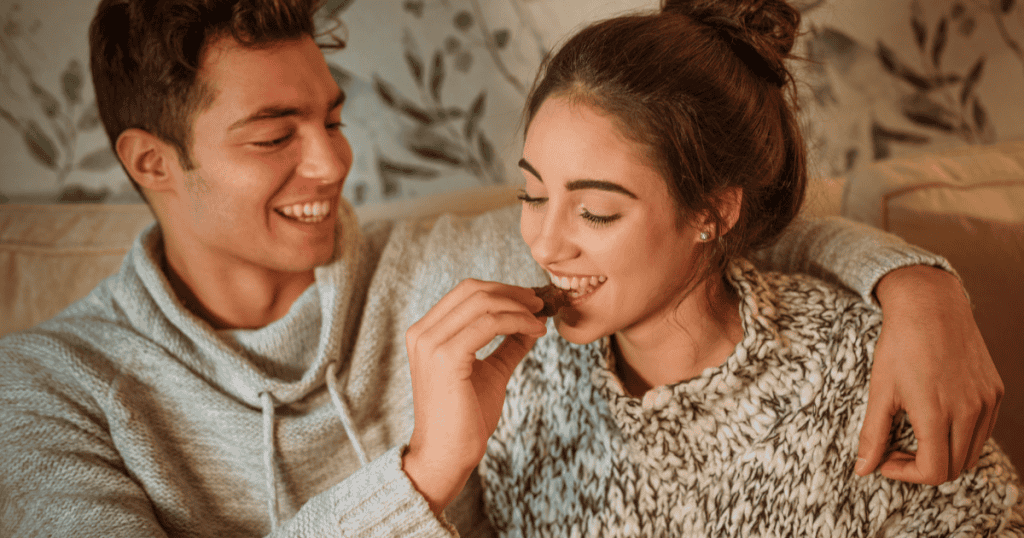 A man playfully feeds chocolate to a smiling woman relaxing beside him on a couch, illustrating how superficial charm and lighthearted affection are often used to maintain disarming control.