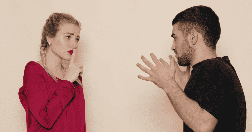A woman holds a finger to her lips in a silencing gesture while a man gestures defensively with both hands, depicting the clash between setting firm personal boundaries and reacting with evasive explanations.
