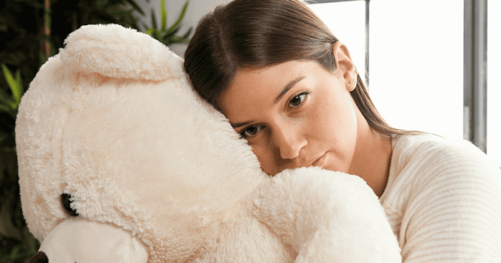 A young woman rests her cheek against a large plush teddy bear with a wistful expression, capturing the quiet search for comfort and safety in a solitary space.