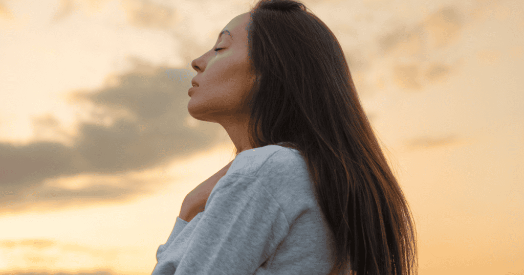 A woman tilts her head back with eyes closed and a hand against her chest under a golden sunset sky, portraying a moment of deep, grounded calm and emotional release.