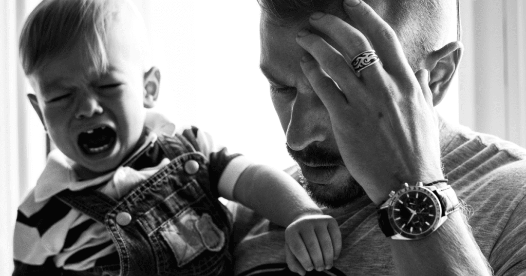 A father rubs his forehead in deep distress while holding a crying toddler in high-contrast black and white, capturing the overwhelming emotional exhaustion that comes from navigating chronic overwhelm.