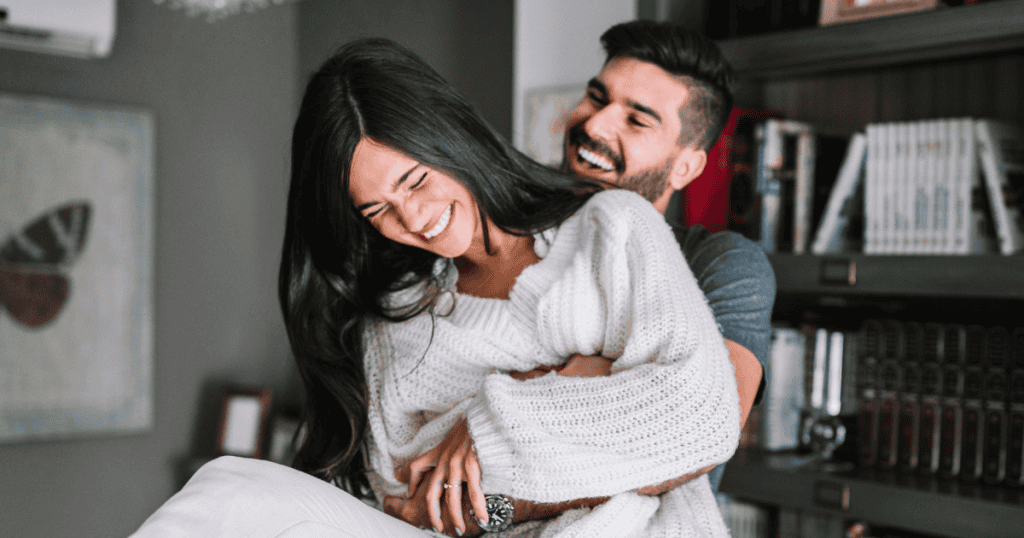 A man playfully hugs a laughing woman from behind in a cozy home setting, showcasing the authentic warmth and spontaneous joy that thrives away from calculated behavior.