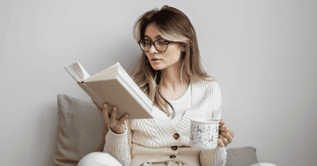 A young woman wearing glasses reads an open book while holding a warm patterned mug, reflecting quiet self-reflection and the comfort of peaceful personal time.