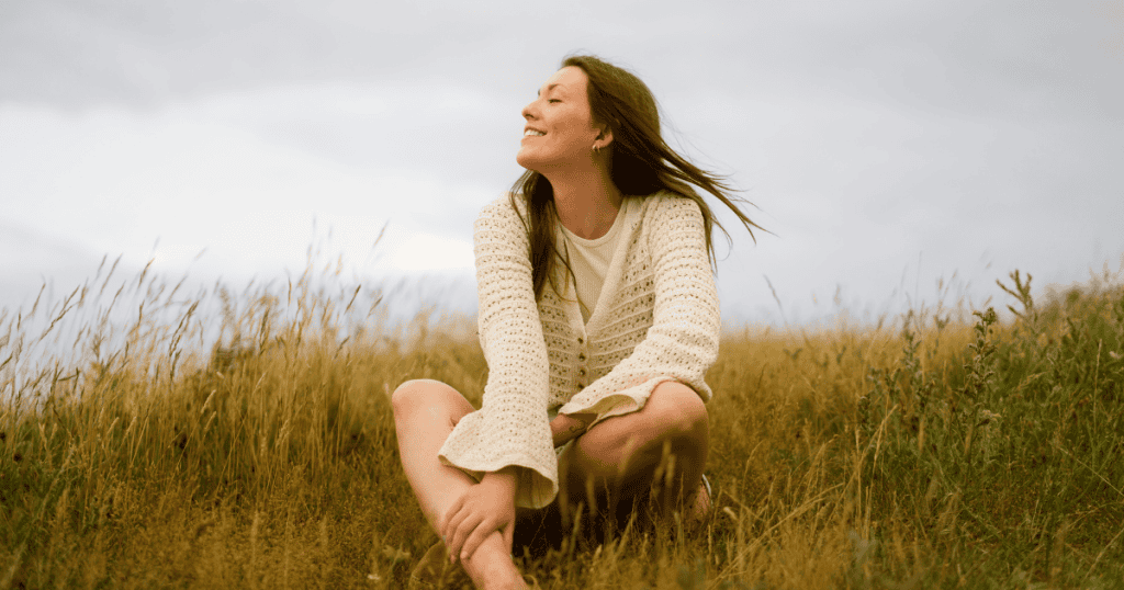 A woman in a knit cardigan sits serenely in an open grassy field with the wind in her hair, embodying the liberating calm found when leaving behind manipulative drama.