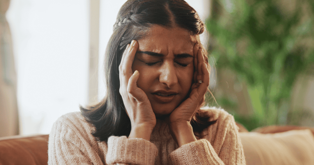 A woman holds both hands to the sides of her head with eyes tightly shut in pain, illustrating the intense physical and emotional toll of overwhelming stress.