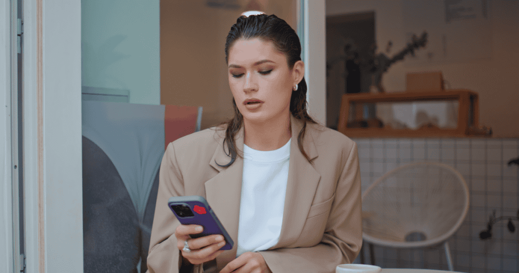 A well-dressed woman sits at an outdoor cafe table looking down at her smartphone with a perplexed, uneasy expression, capturing the sudden jolt of receiving a disruptive, unwelcome message.