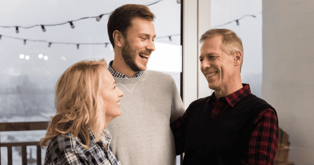 A young man laughs heartily with an older couple by a window, demonstrating how charming, sociable banter is effortlessly used to blend into family spaces.