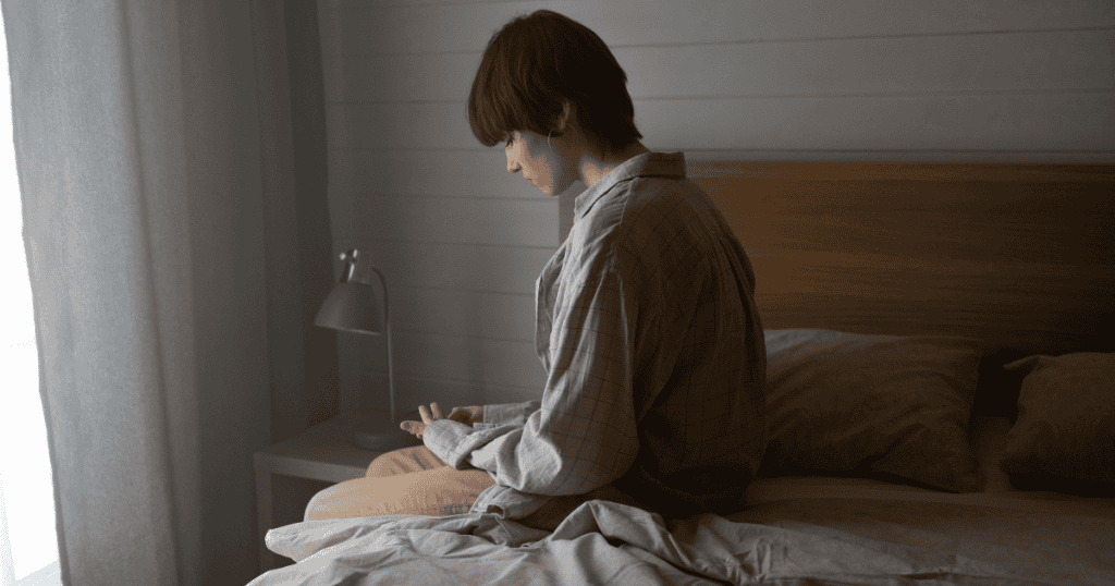 A woman sits alone in profile on the edge of an unmade bed with a downcast posture, depicting the quiet desolation and isolation left behind after repeated emotional strain.