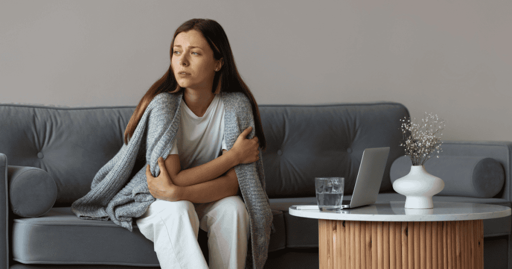 A woman wrapped tightly in a blanket sits on a sofa with an alert, guarded expression, demonstrating that stress responses are natural protective mechanisms rather than personal failure.