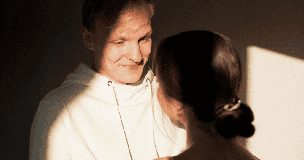 A man smiles down at a woman in dramatic side-lighting while his eyes dart slightly away, capturing the lingering distraction of someone constantly scanning for external validation.