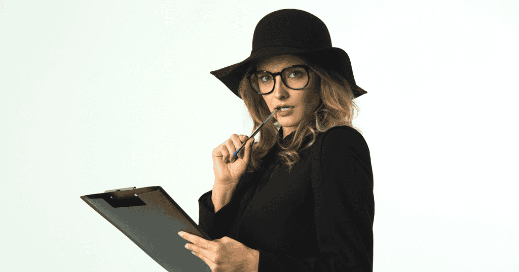 A blonde woman wearing glasses and a wide-brimmed black hat holds a clipboard while biting pensively on her pen, portraying calculated scrutiny and the quiet gathering of behavioral details.