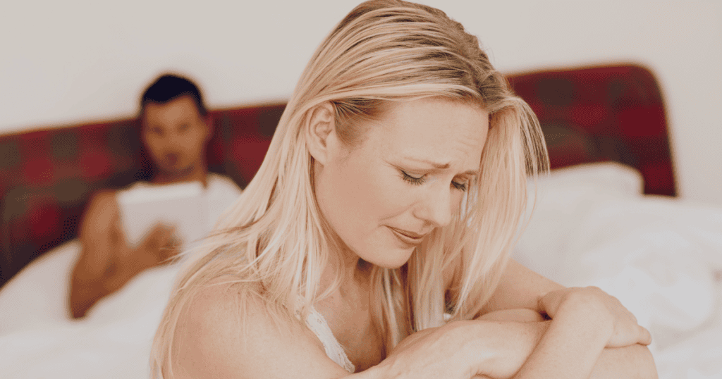 A blonde woman sits weeping at the edge of a bed while her partner remains emotionally detached in the background, portraying the acute isolation of feeling alone within your own marriage.