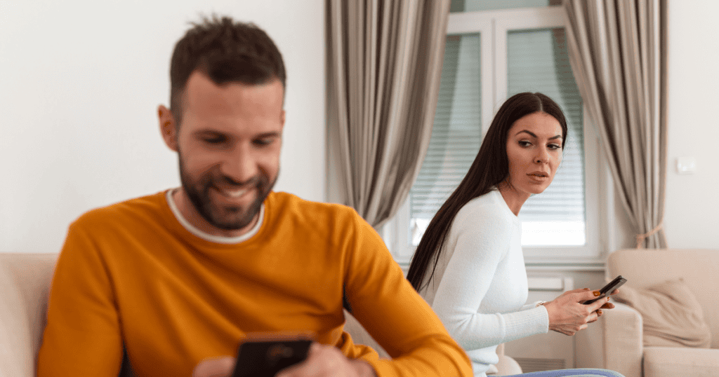 A woman glances suspiciously over her shoulder with her phone in hand while a man smiles at his screen in the foreground, depicting the emotional isolation of being shut out from a partner's private world.