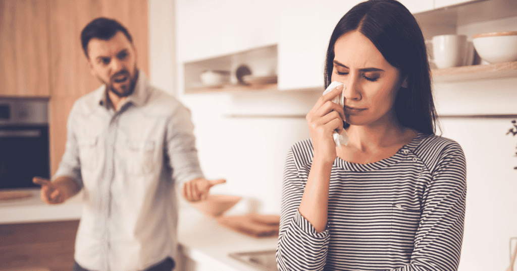 A distressed woman dabs tears with a tissue in the kitchen while a man shouts defensively behind her, illustrating the deep isolation of being mocked for showing natural emotional pain.
